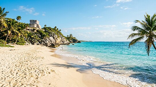 Strand mit weißem Sand, türkisfarbenem Meer, Palmen und einer alten Steinruine auf einem Felsen unter blauem Himmel.